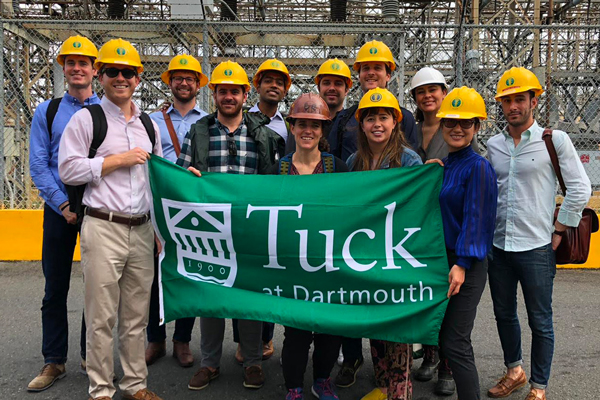 A small group of students wearing hard hats, standing in front of a construction site, holding a Tuck at Dartmouth banner.